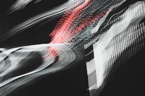 A dynamic and abstract composition with sweeping lines and blurred patterns, featuring interwoven textures of light. Pulsating red and white streaks create a sense of motion against a dark background, evoking a futuristic or digital aesthetic.