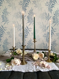 The image features an arrangement of four tall candles in varying colors—white, gray, and green—set in brass candleholders. Surrounding the candles are pale pink and white roses, complemented by scattered petals and greenery. The setup is placed on a soft, white cloth on a dark wooden table. The background is a wallpaper adorned with a subtle fern pattern in soft blue tones.