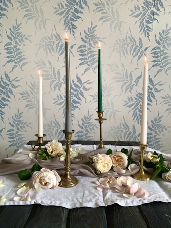 The image features an arrangement of four tall candles in varying colors&mdash;white, gray, and green&mdash;set in brass candleholders. Surrounding the candles are pale pink and white roses, complemented by scattered petals and greenery. The setup is placed on a soft, white cloth on a dark wooden table. The background is a wallpaper adorned with a subtle fern pattern in soft blue tones.
