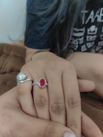 A close-up view of two hands touching each other. One hand is adorned with two rings, one featuring a silver band with a red gemstone and the other a silver design. The individual is wearing a dark-colored shirt with white printed graphics. The background features a soft brown fabric, suggesting a comfortable and warm setting.