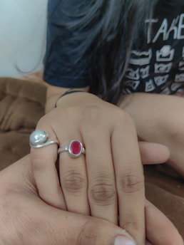 A close-up view of two hands touching each other. One hand is adorned with two rings, one featuring a silver band with a red gemstone and the other a silver design. The individual is wearing a dark-colored shirt with white printed graphics. The background features a soft brown fabric, suggesting a comfortable and warm setting.