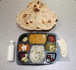 A table setting with a variety of Indian dishes. Three rotis are placed in a basket above a compartmentalized tray filled with different items. The tray includes items like rice garnished with herbs, several curries in varying colors, two round desserts, sliced cucumber and beetroot, and a lemon wedge. A bottle of white drink and a piece of papad are also present.