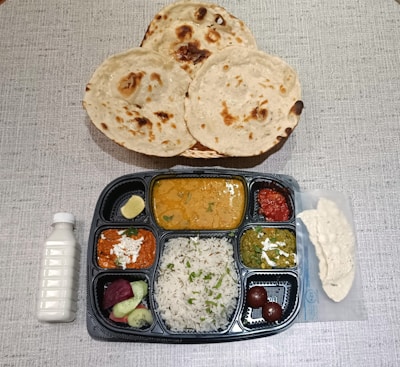 A table setting with a variety of Indian dishes. Three rotis are placed in a basket above a compartmentalized tray filled with different items. The tray includes items like rice garnished with herbs, several curries in varying colors, two round desserts, sliced cucumber and beetroot, and a lemon wedge. A bottle of white drink and a piece of papad are also present.