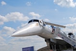 A large, sleek, silver aircraft with a distinctive pointed nose and a set of stairs leading to its entrance is shown against a backdrop of a partly cloudy sky. A few people are visible ascending the stairs towards the plane, which appears to be on display. Various logos and lettering are visible on the fuselage, indicating the aircraft's affiliations and purpose.