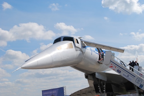 A large, sleek, silver aircraft with a distinctive pointed nose and a set of stairs leading to its entrance is shown against a backdrop of a partly cloudy sky. A few people are visible ascending the stairs towards the plane, which appears to be on display. Various logos and lettering are visible on the fuselage, indicating the aircraft's affiliations and purpose.