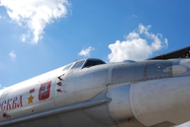 A close-up view of the fuselage of a military aircraft with visible insignia, including a red shield with a detailed emblem and Cyrillic writing. The metal surface is shiny and reflective under the bright daylight, with a clear blue sky and scattered white clouds in the background.