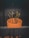 A cactus with multiple arms is planted in a large, round, orange pot. The background features a dark wall, creating a stark contrast with the bright color of the pot. The setting appears to be indoors, possibly a decorative arrangement.