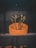 A cactus with multiple arms is planted in a large, round, orange pot. The background features a dark wall, creating a stark contrast with the bright color of the pot. The setting appears to be indoors, possibly a decorative arrangement.