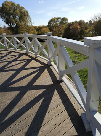 A wooden deck with white railings casts shadows on the planks. In the background, trees with autumn foliage are visible under a clear blue sky.