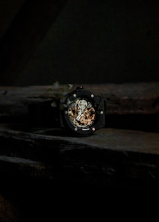 A close-up view of a black watch with an intricately designed open-face dial displaying its mechanical components. The background is dark, making the watch the focal point.