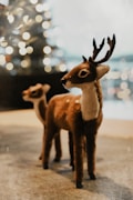 Two plush deer figurines stand on a surface, with soft, out-of-focus lights in the background suggesting a festive or cozy atmosphere. The deer are detailed with realistic textures, emphasizing a warm, handcrafted quality.