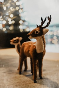 Two plush deer figurines stand on a surface, with soft, out-of-focus lights in the background suggesting a festive or cozy atmosphere. The deer are detailed with realistic textures, emphasizing a warm, handcrafted quality.