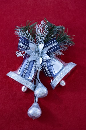 Shiny silver bells decorated with a blue ribbon and floral elements are set against a rich red background. The composition includes shimmering beads and pine foliage for a festive appearance.