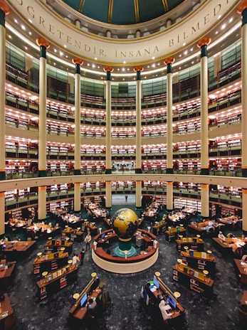 A large, multi-tiered library with a central globe installation. The library is circular in design, with several levels of bookshelves stacked one above the other. There are numerous desks equipped with computers and stacks of books, occupied by readers and researchers. Tall columns support the structure, and there is ornate decoration with lights on each level. The phrase 'Öğretendir, insana bilmedim' is inscribed at the dome of the structure.
