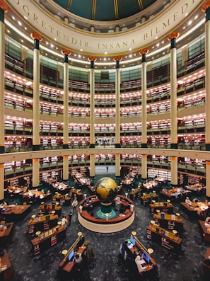 A large, multi-tiered library with a central globe installation. The library is circular in design, with several levels of bookshelves stacked one above the other. There are numerous desks equipped with computers and stacks of books, occupied by readers and researchers. Tall columns support the structure, and there is ornate decoration with lights on each level. The phrase 'Öğretendir, insana bilmedim' is inscribed at the dome of the structure.