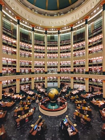 A large, multi-tiered library with a central globe installation. The library is circular in design, with several levels of bookshelves stacked one above the other. There are numerous desks equipped with computers and stacks of books, occupied by readers and researchers. Tall columns support the structure, and there is ornate decoration with lights on each level. The phrase '&Ouml;ğretendir, insana bilmedim' is inscribed at the dome of the structure.
