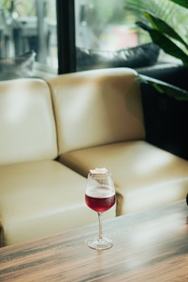 A glass of red wine with a garnish on top sits on a wooden table in front of a beige sofa. The background includes a window with a view of greenery, suggesting a relaxed indoor setting.
