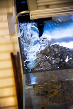 Water flows into an aquarium, creating bubbles on the surface. The tank contains a textured object or coral visible through the clear water, and there are reflections of light creating a calming visual effect.