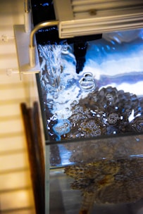 Water flows into an aquarium, creating bubbles on the surface. The tank contains a textured object or coral visible through the clear water, and there are reflections of light creating a calming visual effect.
