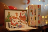 A Christmas-themed pop-up card featuring Santa Claus, a reindeer, a Christmas tree, and various holiday elements is displayed prominently. Next to it is a wrapped gift box with colorful paper showcasing cartoon-like patterns. Warm lighting and blurred background create a festive ambiance.