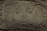 A bas-relief sculpture featuring a central figure with arms raised, surrounded by stylized animal figures resembling lions or other mythical creatures. The artwork is intricate, with swirling patterns and foliage motifs.
