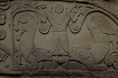 A bas-relief sculpture featuring a central figure with arms raised, surrounded by stylized animal figures resembling lions or other mythical creatures. The artwork is intricate, with swirling patterns and foliage motifs.
