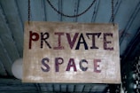 A rectangular wooden sign hangs from a chain. It has the words 'PRIVATE SPACE' painted in a mix of red, black, and brown on a distressed beige background. The sign has a rustic appearance and is set against a background of teal wooden boards.