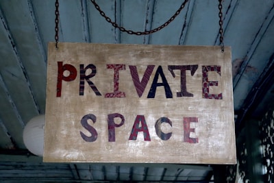 A rectangular wooden sign hangs from a chain. It has the words 'PRIVATE SPACE' painted in a mix of red, black, and brown on a distressed beige background. The sign has a rustic appearance and is set against a background of teal wooden boards.