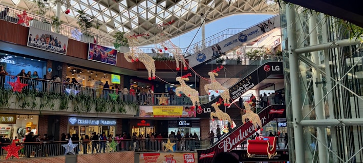 A multi-level shopping mall decorated festively with red and gold reindeer and stars hanging from the ceiling. Various shop fronts, such as Puma, Shree Balaji Diamond, and others, are visible. People are shopping and walking along the balconies. There are advertisements for Budweiser and LG.