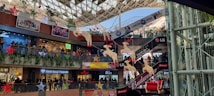 A multi-level shopping mall decorated festively with red and gold reindeer and stars hanging from the ceiling. Various shop fronts, such as Puma, Shree Balaji Diamond, and others, are visible. People are shopping and walking along the balconies. There are advertisements for Budweiser and LG.