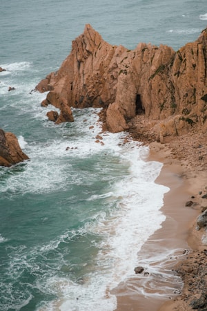 A rugged coastline features dramatic cliffs jutting into the ocean, with waves crashing against the rocky shore. The sea is a vibrant teal, and the sandy beach is lined with small rocks and pebbles. The landscape suggests a natural and untouched environment.