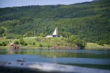 A serene landscape features a hillside with lush greenery and dense forests. A modern, triangular-roofed building sits prominently on the hill. In the foreground, a calm body of water reflects the surrounding scenery, and two small red cabins can be seen near the water's edge.