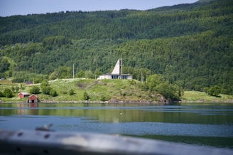 A serene landscape features a hillside with lush greenery and dense forests. A modern, triangular-roofed building sits prominently on the hill. In the foreground, a calm body of water reflects the surrounding scenery, and two small red cabins can be seen near the water's edge.