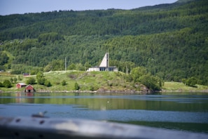 A serene landscape features a hillside with lush greenery and dense forests. A modern, triangular-roofed building sits prominently on the hill. In the foreground, a calm body of water reflects the surrounding scenery, and two small red cabins can be seen near the water's edge.
