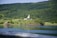 A serene landscape features a hillside with lush greenery and dense forests. A modern, triangular-roofed building sits prominently on the hill. In the foreground, a calm body of water reflects the surrounding scenery, and two small red cabins can be seen near the water's edge.