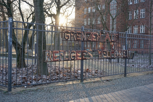 A metal fence adorned with a sign reading 'GRENZEN AUF REFUGEES WELCOME' in uneven letters. Behind the fence, leafless trees and an old brick building with multiple windows are partially illuminated by the setting or rising sun, casting a warm light and long shadows on the frosted ground and cobblestone path.