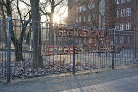 A metal fence adorned with a sign reading 'GRENZEN AUF REFUGEES WELCOME' in uneven letters. Behind the fence, leafless trees and an old brick building with multiple windows are partially illuminated by the setting or rising sun, casting a warm light and long shadows on the frosted ground and cobblestone path.