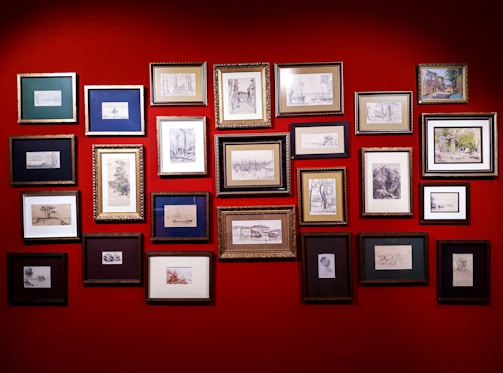 A collection of framed artworks are arranged on a vibrant red wall. The frames vary in size and color, displaying a variety of sketches and paintings, predominantly in black and white with a few color highlights. The artworks seem to include landscapes, portraits, and abstract designs.