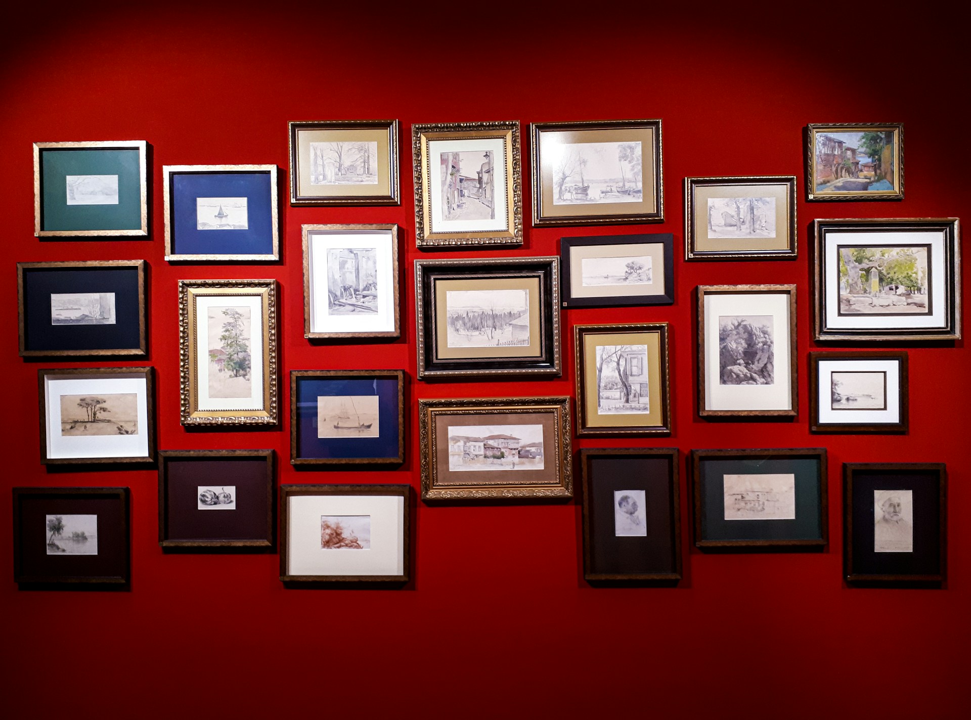A collection of framed artworks are arranged on a vibrant red wall. The frames vary in size and color, displaying a variety of sketches and paintings, predominantly in black and white with a few color highlights. The artworks seem to include landscapes, portraits, and abstract designs.