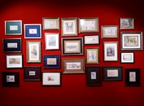 A collection of framed artworks are arranged on a vibrant red wall. The frames vary in size and color, displaying a variety of sketches and paintings, predominantly in black and white with a few color highlights. The artworks seem to include landscapes, portraits, and abstract designs.