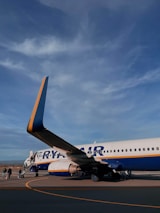A commercial airplane on the tarmac with passengers boarding via an outdoor staircase. The aircraft is marked with blue and yellow colors and the branding is partly visible. The sky above is clear with some clouds visible.