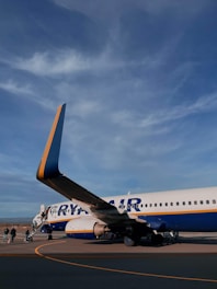 A commercial airplane on the tarmac with passengers boarding via an outdoor staircase. The aircraft is marked with blue and yellow colors and the branding is partly visible. The sky above is clear with some clouds visible.