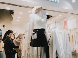 A clothing store scene with a mannequin dressed in a white sweater, black skirt, and a pearl bag. The store is well-lit, displaying neatly arranged clothing racks filled with white and beige garments. Two customers are browsing the selection, inspecting items. The atmosphere is modern and inviting.