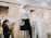 A clothing store scene with a mannequin dressed in a white sweater, black skirt, and a pearl bag. The store is well-lit, displaying neatly arranged clothing racks filled with white and beige garments. Two customers are browsing the selection, inspecting items. The atmosphere is modern and inviting.