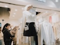A clothing store scene with a mannequin dressed in a white sweater, black skirt, and a pearl bag. The store is well-lit, displaying neatly arranged clothing racks filled with white and beige garments. Two customers are browsing the selection, inspecting items. The atmosphere is modern and inviting.