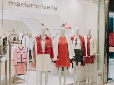 A clothing store display featuring several mannequins dressed in stylish outfits. The mannequins are adorned in red and white clothing, including dresses and blazers. Behind them, more clothes are visible on hangers, indicating a chic and fashionable boutique. The store's name, 'mademoiselle,' is visible above the display.