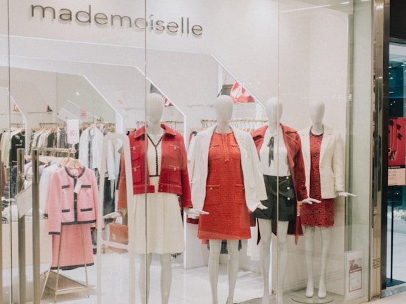 A clothing store display featuring several mannequins dressed in stylish outfits. The mannequins are adorned in red and white clothing, including dresses and blazers. Behind them, more clothes are visible on hangers, indicating a chic and fashionable boutique. The store's name, 'mademoiselle,' is visible above the display.