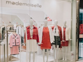 A clothing store display featuring several mannequins dressed in stylish outfits. The mannequins are adorned in red and white clothing, including dresses and blazers. Behind them, more clothes are visible on hangers, indicating a chic and fashionable boutique. The store's name, 'mademoiselle,' is visible above the display.