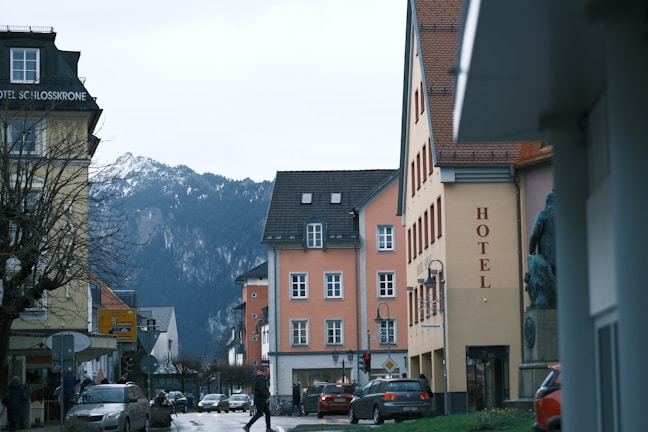 A street scene featuring several buildings with colorful facades, including a yellow hotel with a red sign. Cars are parked along the road, and a few pedestrians are walking. In the background, snow-capped mountains provide a dramatic landscape.