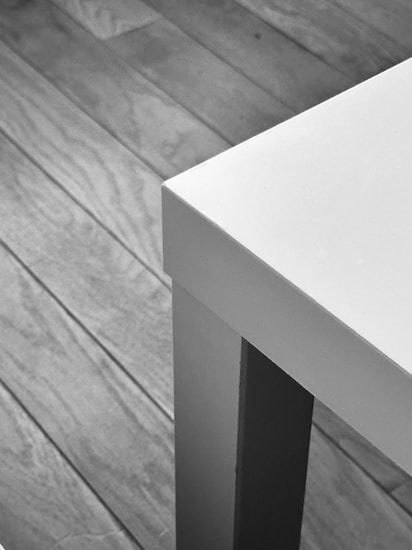 A black and white image showcasing a wooden floor and the corner of a modern, rectangular white table with sleek lines.